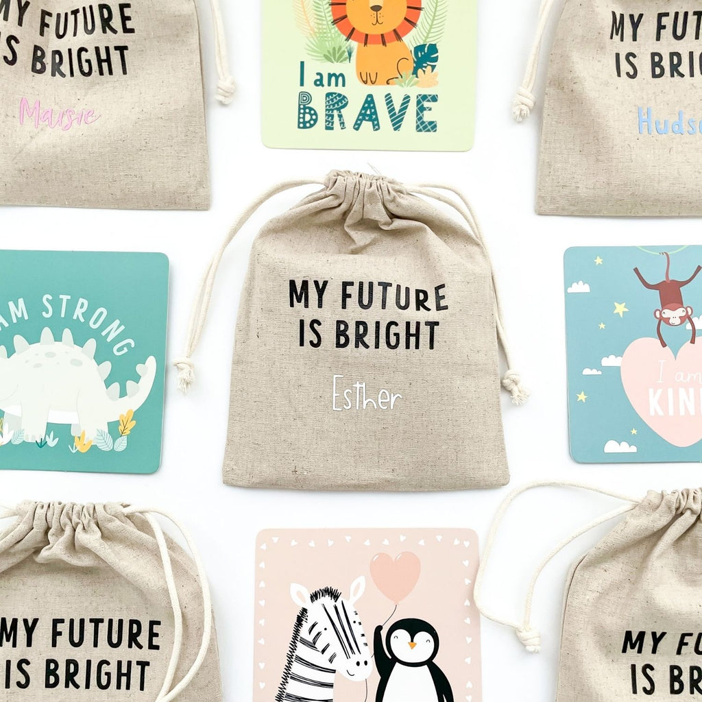 Flat lay showing a selection of Children's Affirmation Cards including, "I am Brave, I am Strong, I am Kind. Also displaying the personalized fabric keepsake bag. 